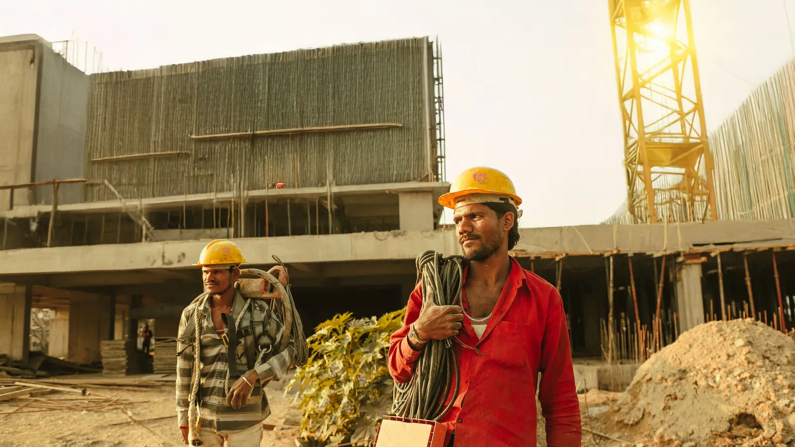 Two workers in hard hats carrying cable and tools across a building site